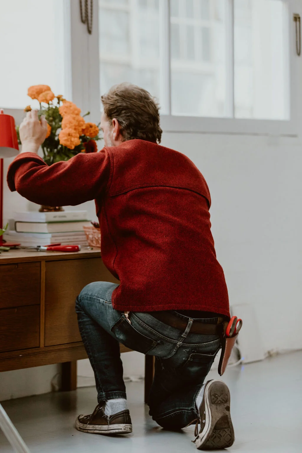 A man kneeling on one knee arranging orange flowers on a desk with books and scissors, in a room with a window.