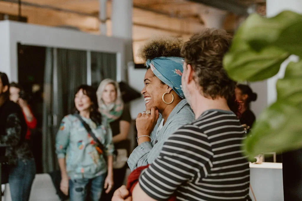 People in a social gathering, smiling and chatting, with a woman wearing a blue headscarf and hoop earrings in focus.
