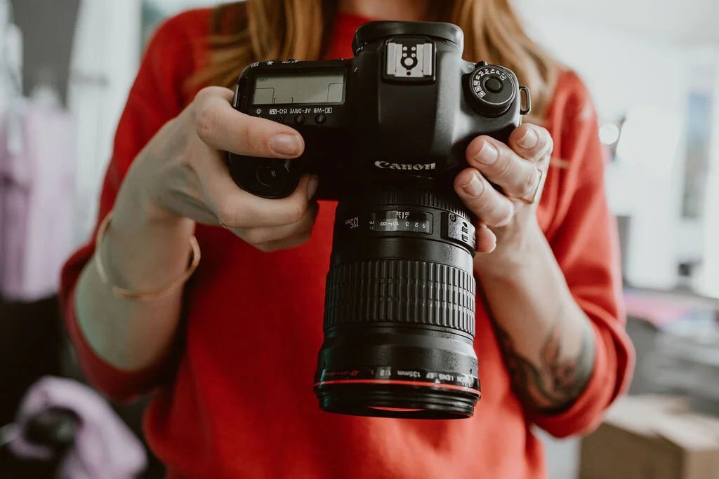 Person holding a Canon DSLR camera with a zoom lens, wearing a red shirt, in an indoor setting.