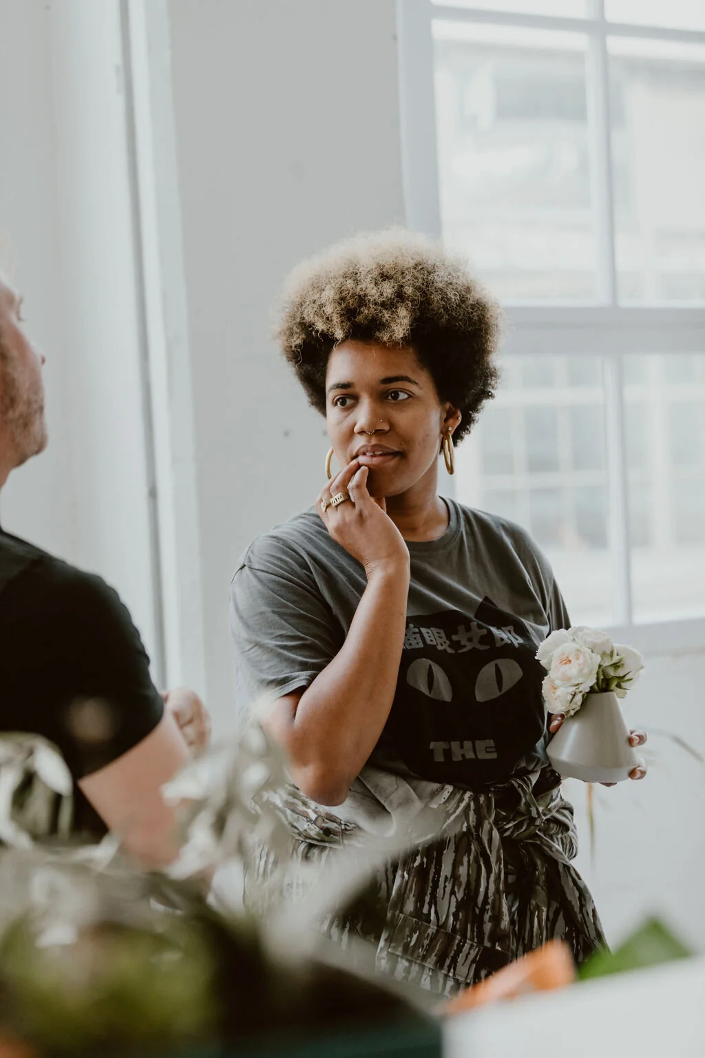 A woman with a curly two-tone hairstyle, wearing a gray t-shirt and patterned skirt, holding a small vase with flowers, engaged in conversation with a man in a black t-shirt.