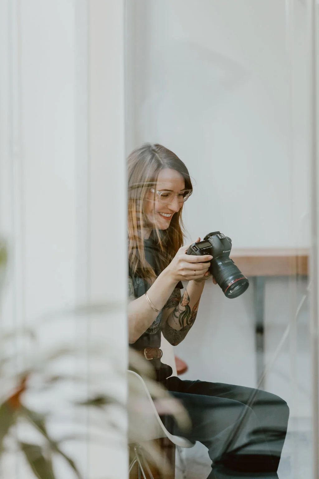 A woman with glasses and tattoos on her arm, sitting on a chair and smiling while looking at a camera she is holding.