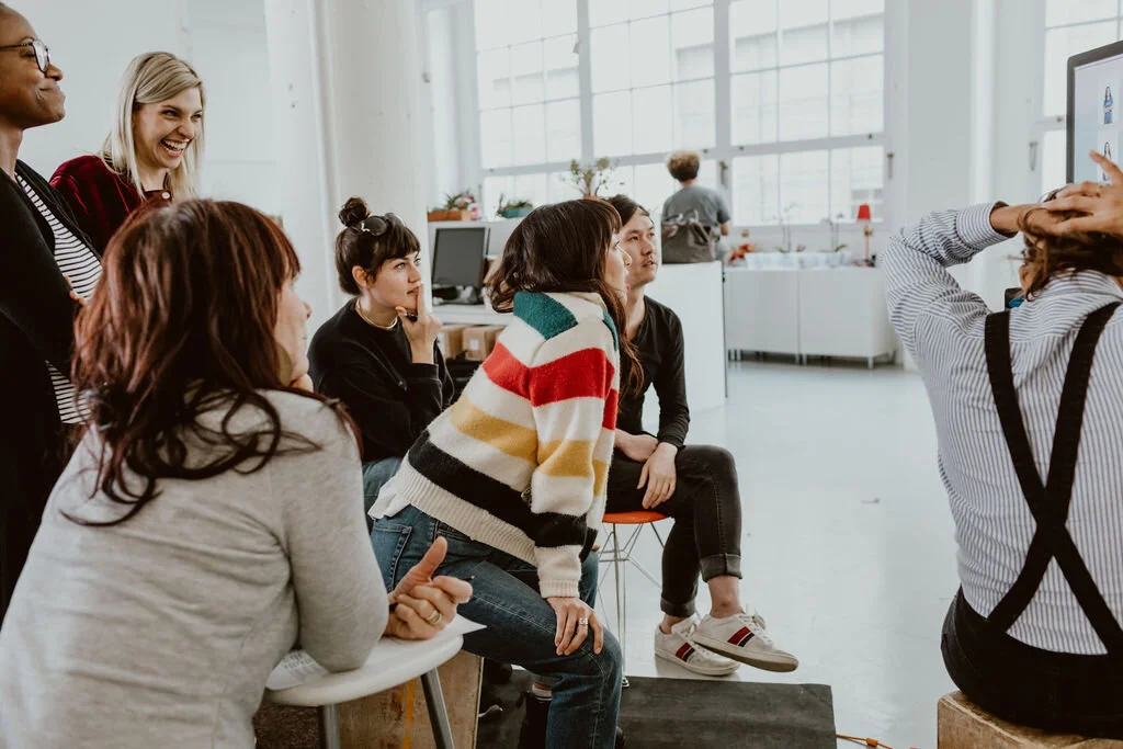 Group of people in a bright, modern room during a photo shoot or class, with some sitting and others standing, smiling and posing for the camera.