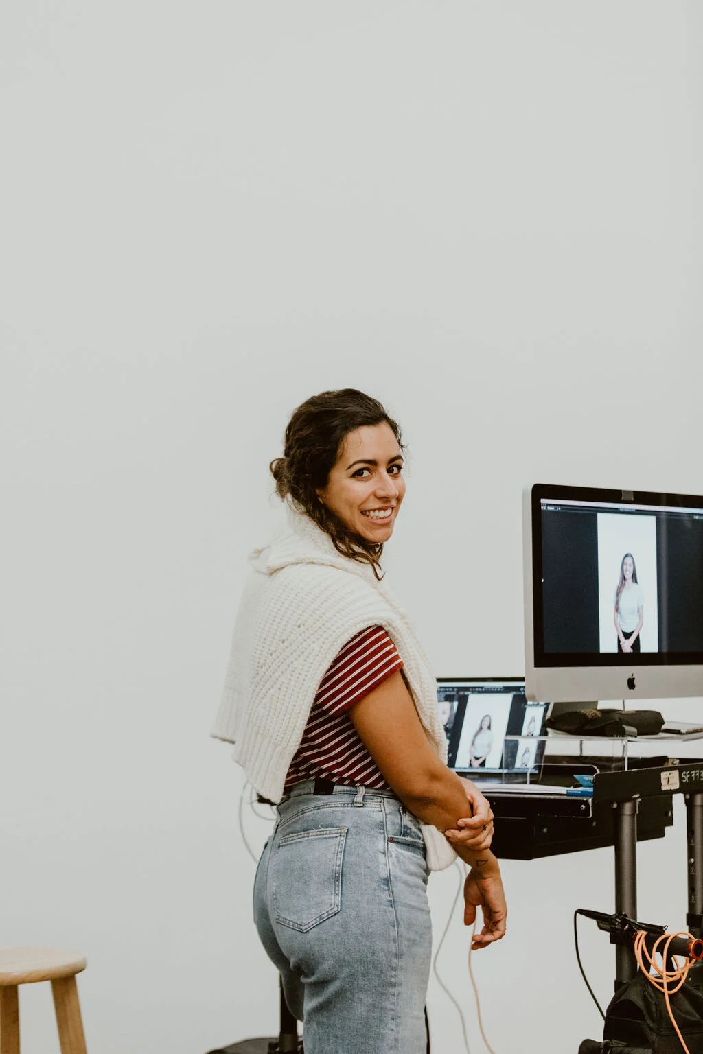A woman with curly hair smiling at the camera while in a photo studio, standing in front of a computer screen displaying a photo of herself, with additional photo editing equipment and a stool nearby.