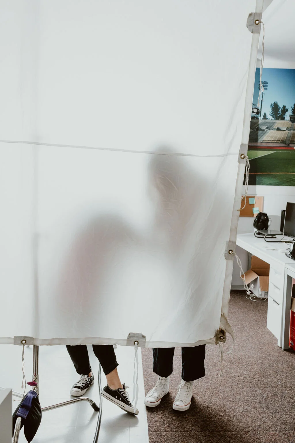 Two people standing behind a bright reflective backdrop, with only their legs visible, in a room with a computer and a large sports field picture on the wall.