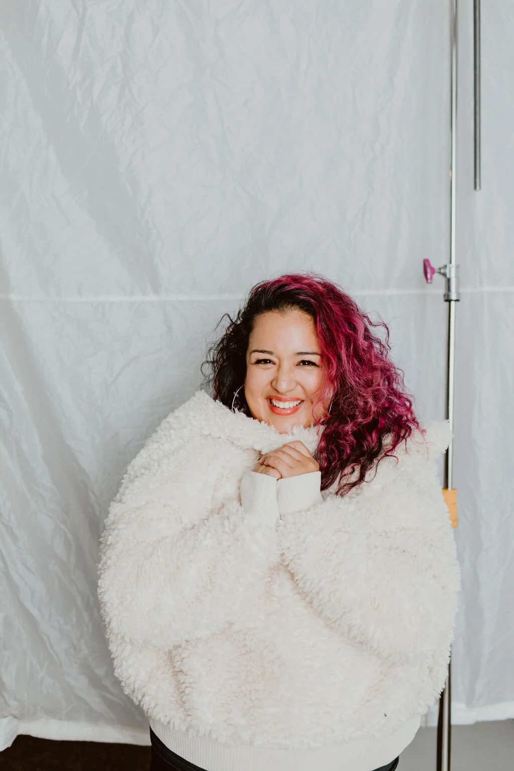 A woman with curly pink hair smiling and hugging herself wrapped in a white fuzzy blanket.