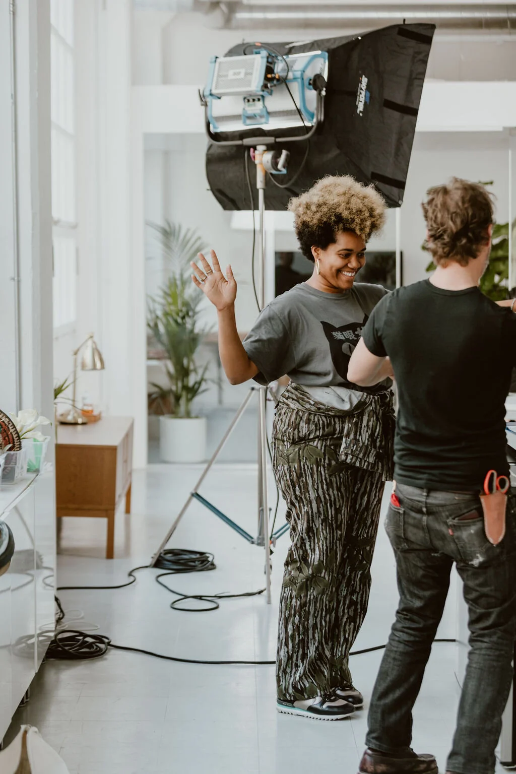 A woman with curly blonde hair smiling and gesturing with her hand during a photo shoot, standing next to another person in a black shirt, in a well-lit indoor studio with lighting equipment and plants.