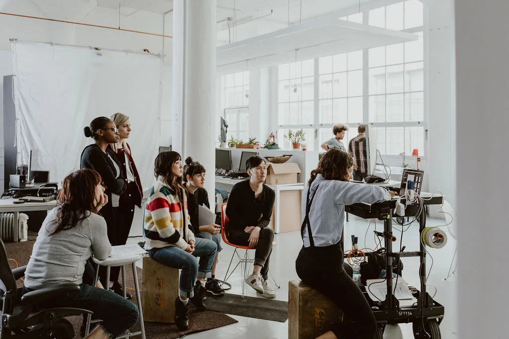 Group of people observing a woman working with camera equipment in a bright, industrial-style office or studio.