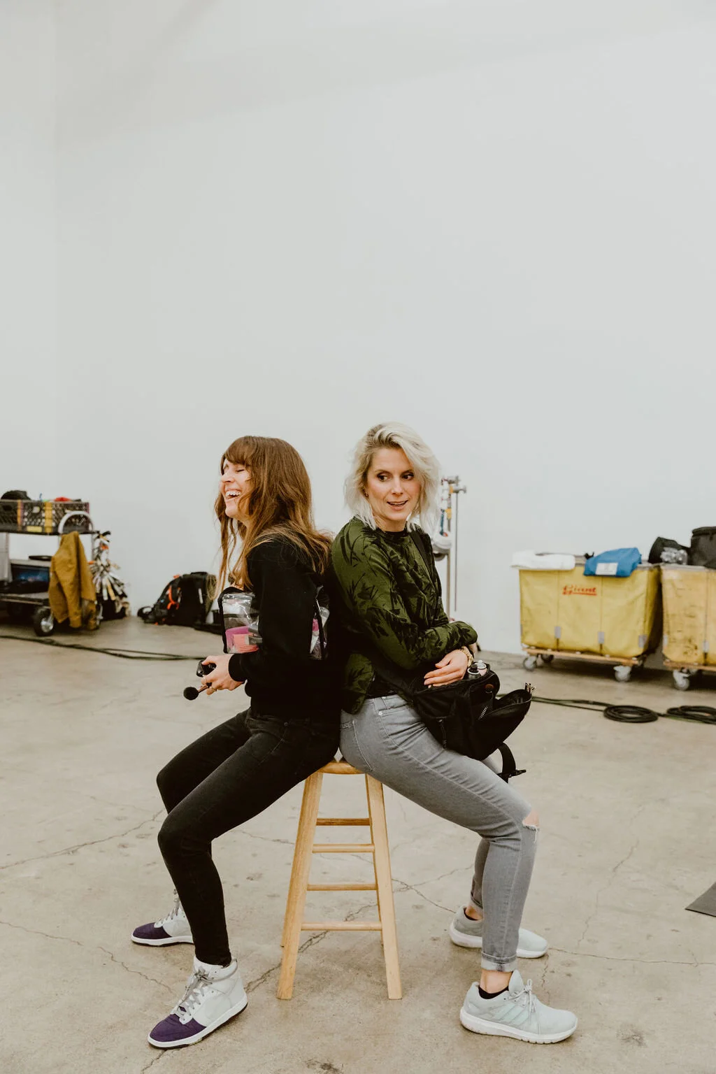 Two women sitting back-to-back on a wooden stool in a large, white room, smiling and laughing. The woman on the left has long, brown hair and is wearing a black sweater and black jeans. The woman on the right has short, blonde hair and is wearing a g
