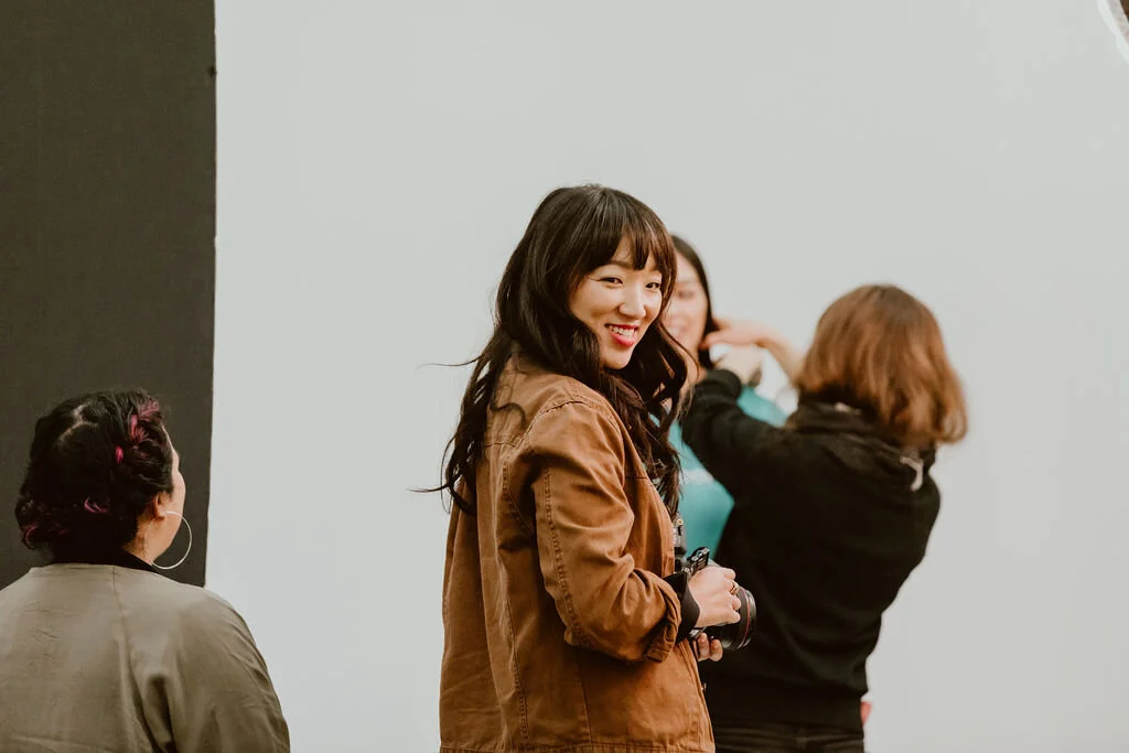 Young woman with long dark hair smiling while holding a camera, standing in front of a plain light gray wall with other people in the background.