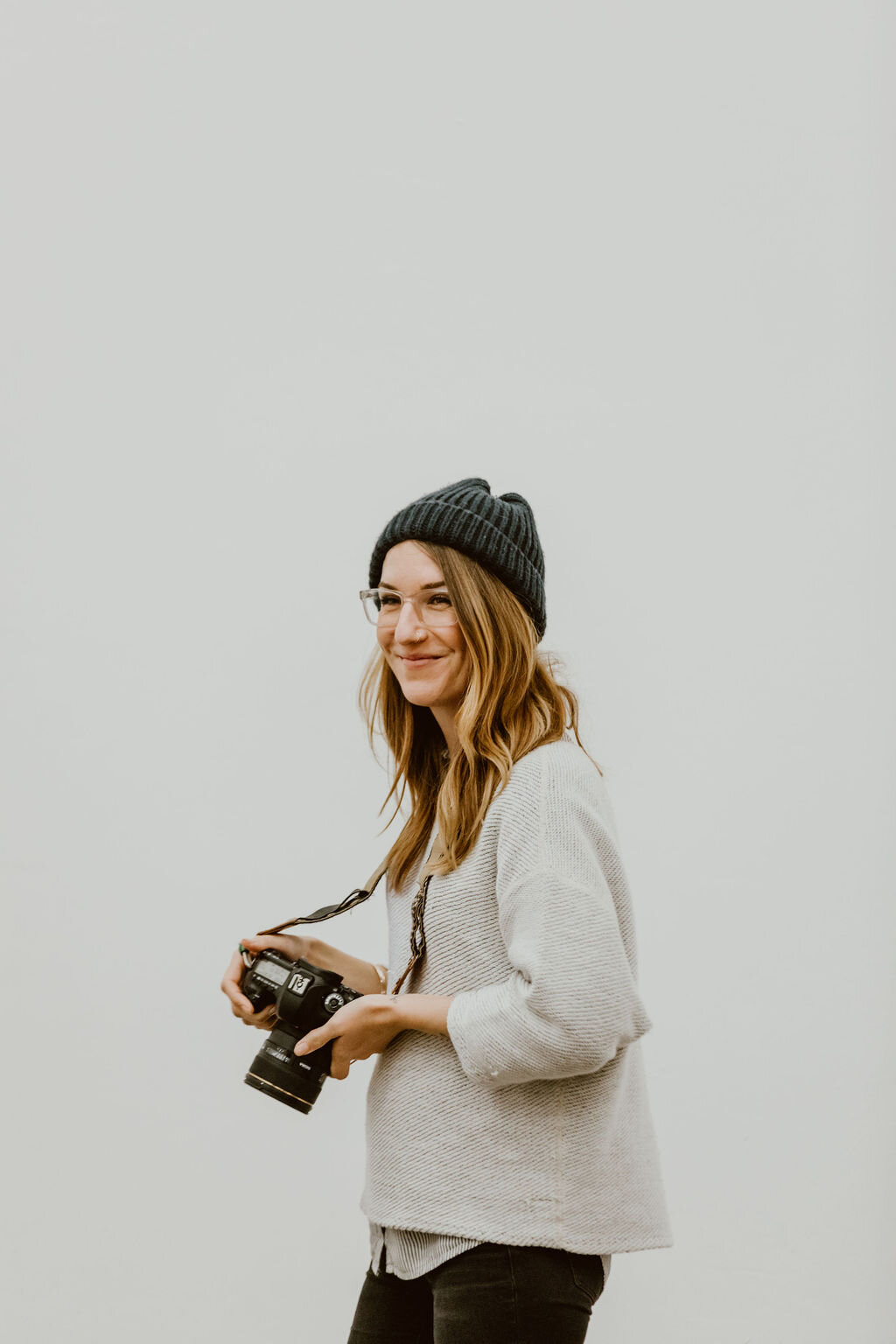 A woman with long wavy hair, wearing a gray knit beanie, glasses, a gray sweater, and black pants, holding a camera, standing against a plain white wall.