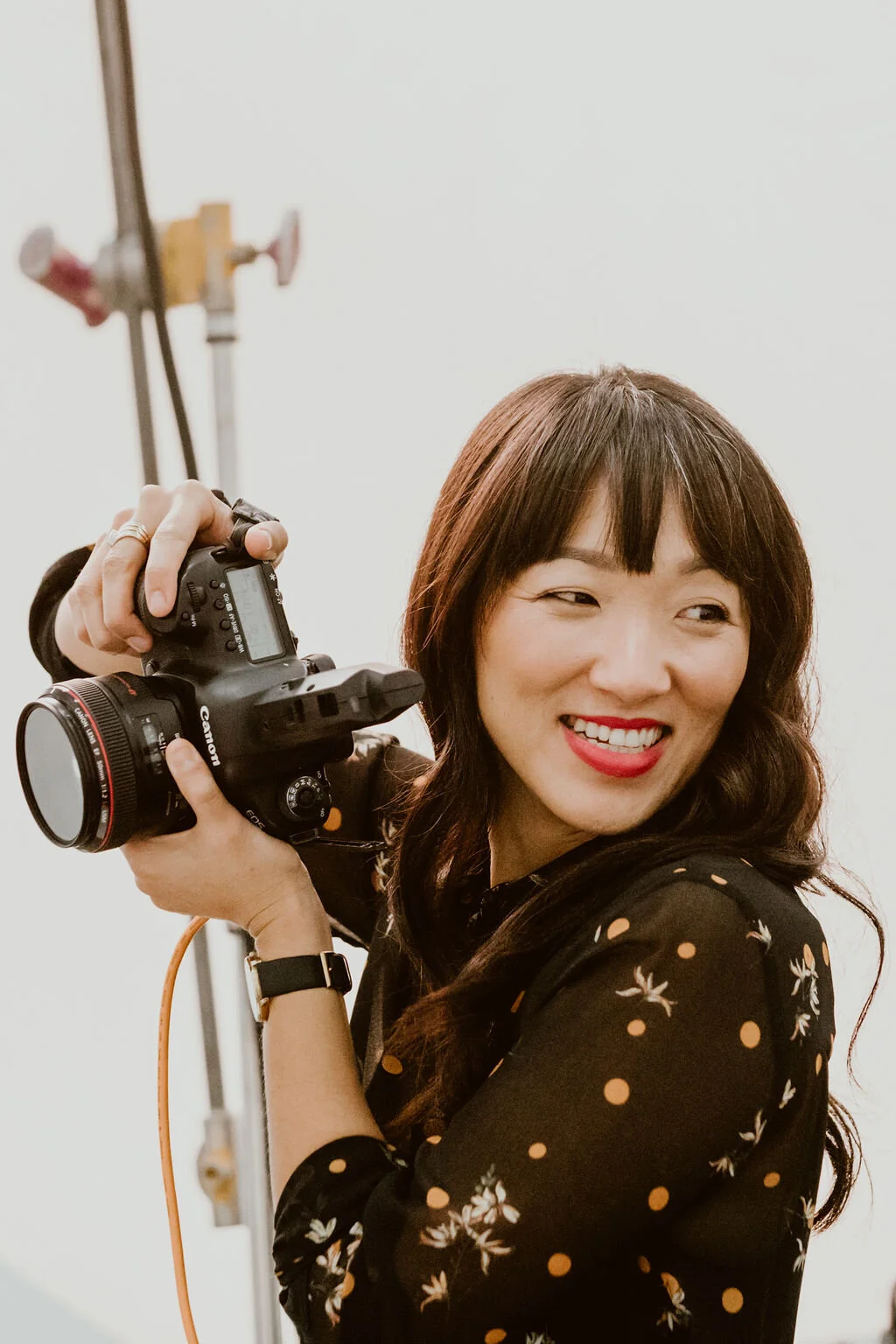 A woman with dark brown hair, wearing a black dress with a floral and polka dot pattern, holding a camera, smiling, and looking to the side in a studio setting.