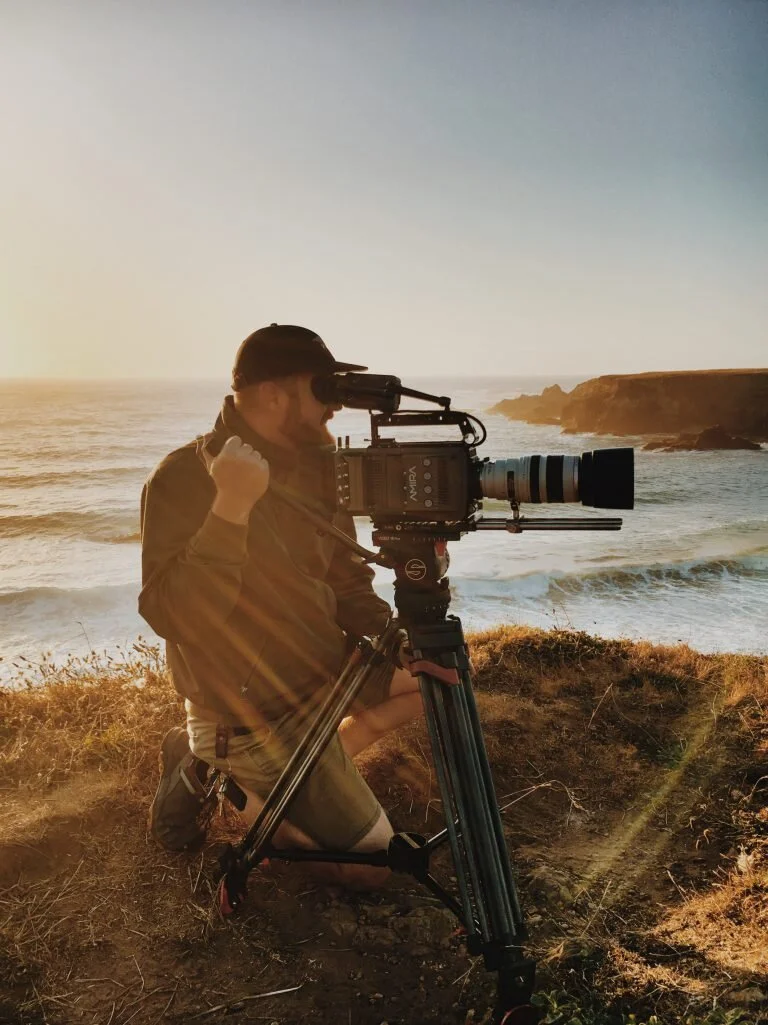 Person operating a camera on a tripod at a coastal location during sunset.