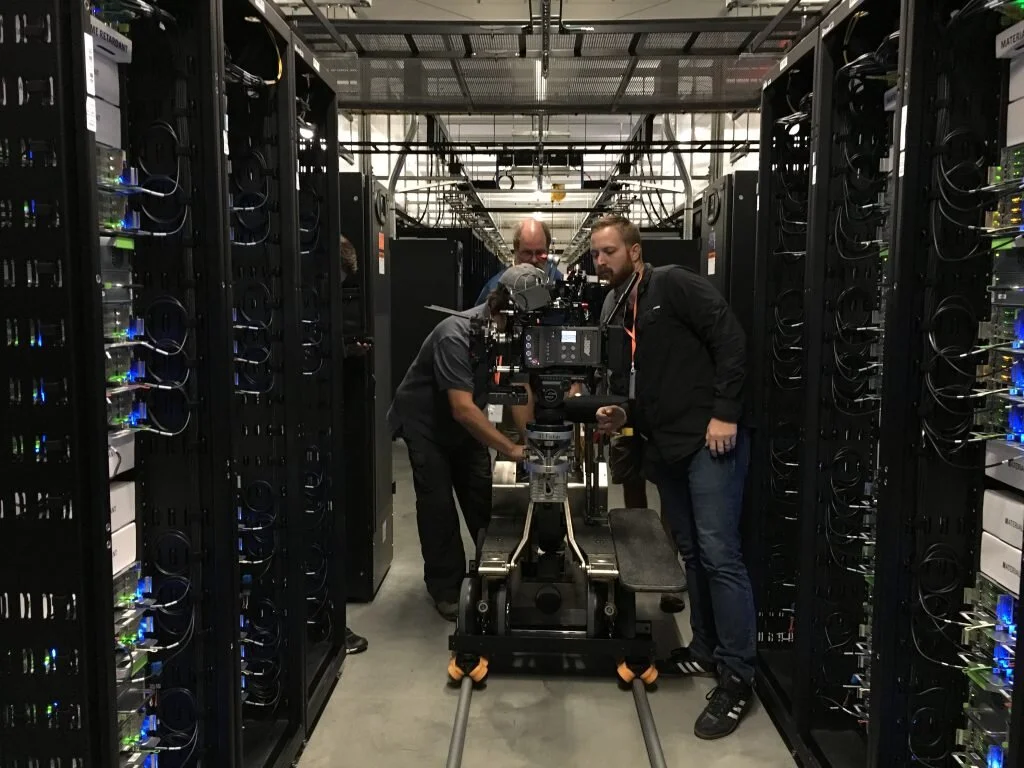 Three men working with a camera on a dolly in a server room with rows of computer servers.
