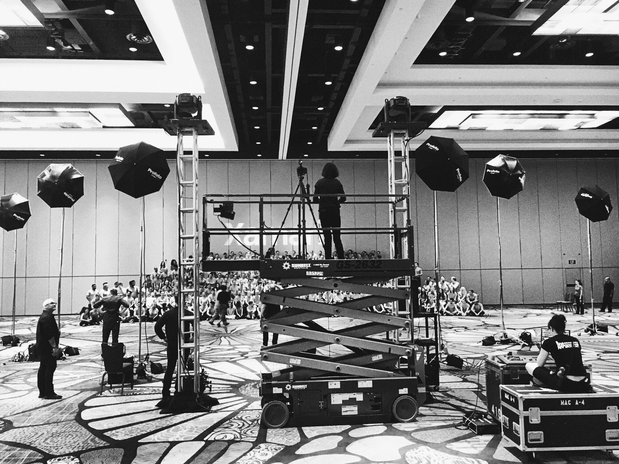 A stage setup in a large conference hall with lifts, lighting equipment, and a person organizing the equipment. Audience seating and people preparing are visible in the background.
