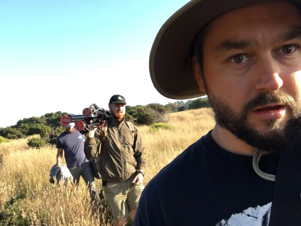 A group of three men walking through a grassy field on a sunny day, with one man taking a selfie in the foreground and the others walking behind him, one carrying a tripod.