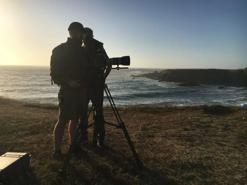Two people, one with a large camera on a tripod, standing near the ocean at sunset or sunrise.