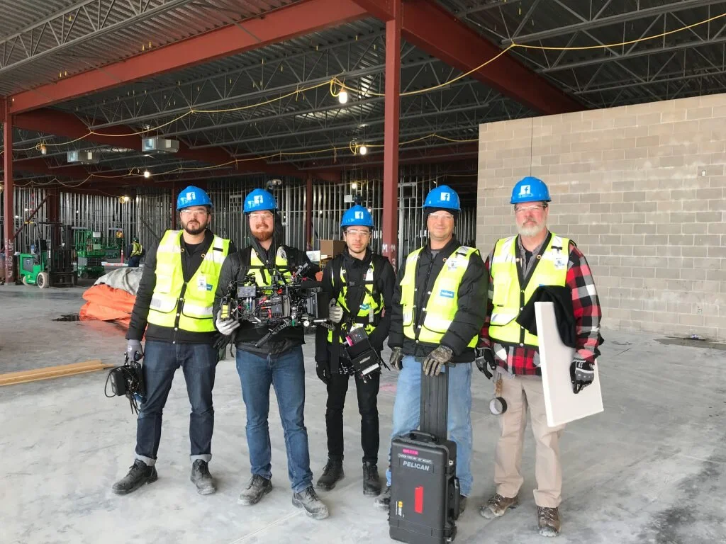 Group of five construction workers on a building site, wearing safety vests and blue hard hats, with some holding and carrying equipment.