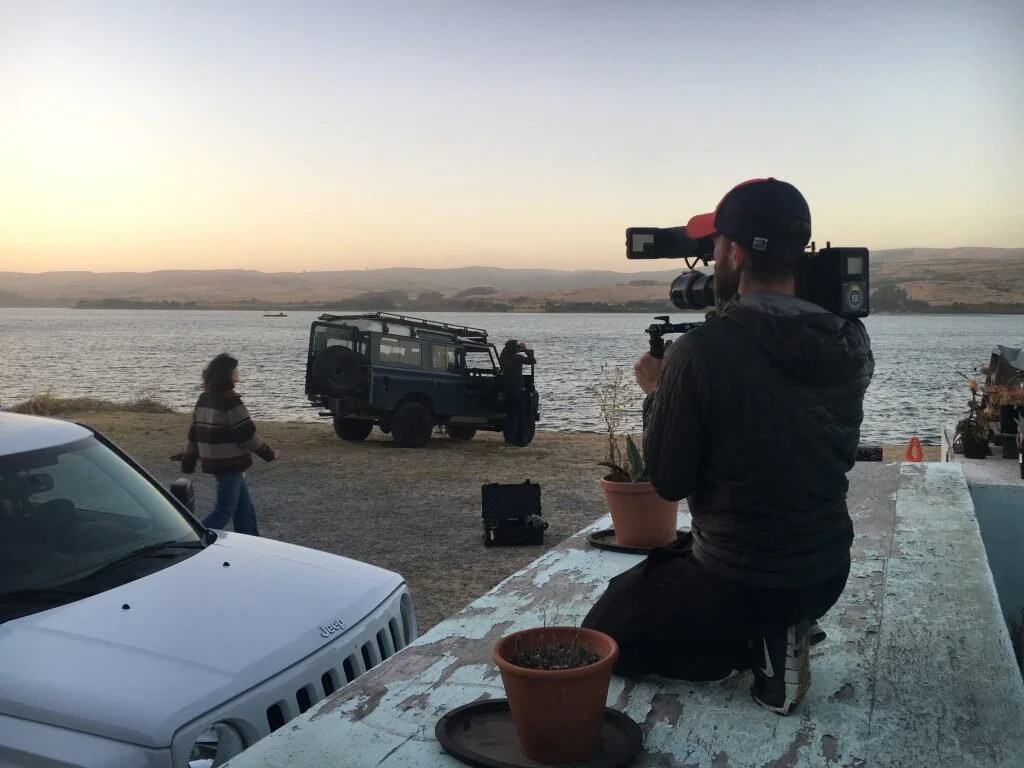A man filming with a large camera on a tripod by the water at sunset, a woman walking nearby, and parked vehicles including a Jeep near a concrete ledge with potted plants.