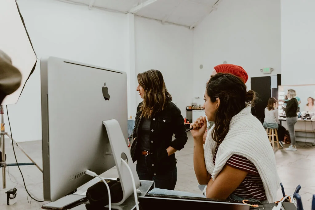 Three women working together in a creative studio, with one looking at the computer screen and the other two observing and discussing.