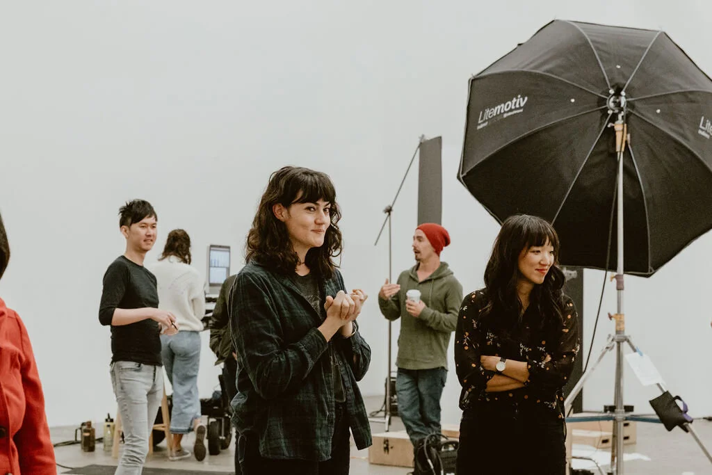 People standing and talking indoors with a large umbrella and a projector in the background.