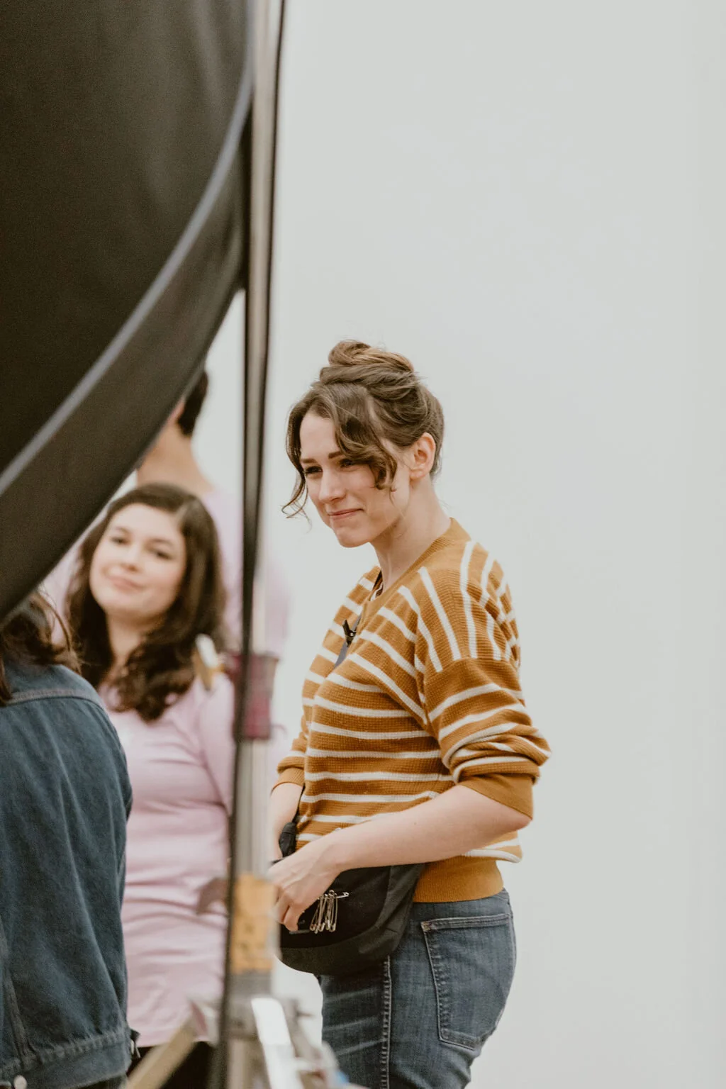 Two women standing near a grand piano inside a bright room. One woman, in the foreground, is wearing a striped brown and white top and jeans, smiling softly. The other woman, in the background, is wearing a pink top and also smiling.