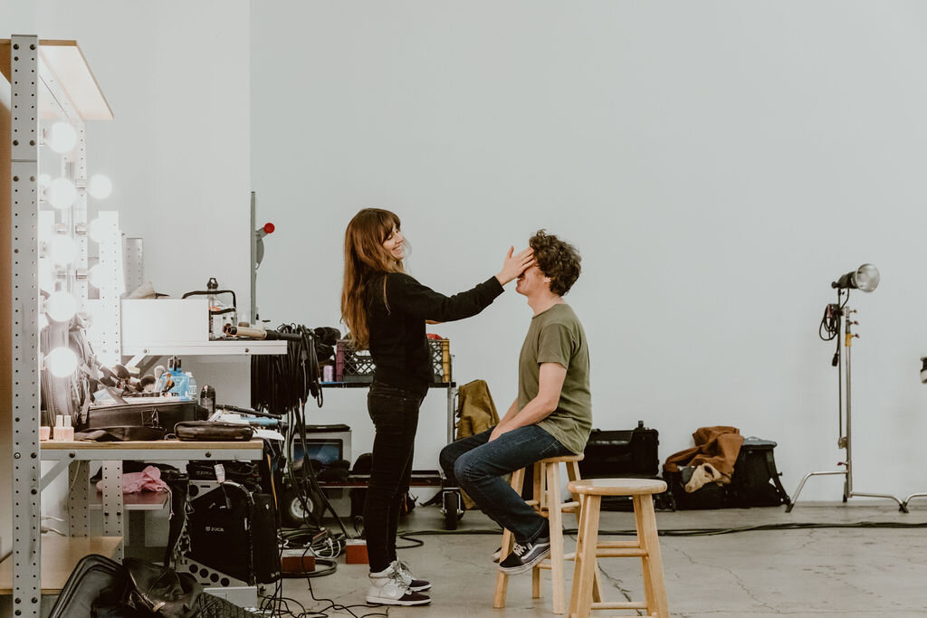 Makeup artist applying makeup to a young man sitting on a stool in a photography studio.