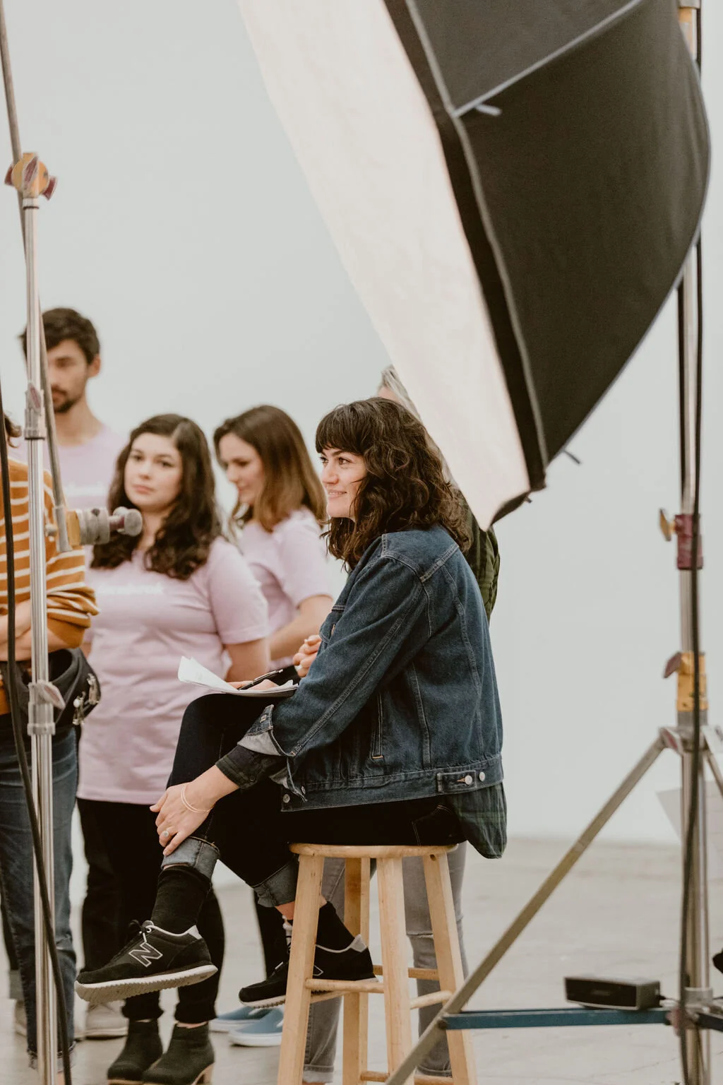 A woman with dark curly hair sitting on a wooden stool wearing a denim jacket, black shoes, and black jeans, smiling and looking to the side during a photo shoot with studio equipment around.