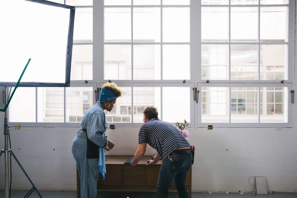 Two people are standing near a wooden table in front of large windows, in a photo studio or office space. One person, wearing a striped shirt, is leaning on the table, while the other, wearing a striped jumpsuit and a headscarf, is standing and looki