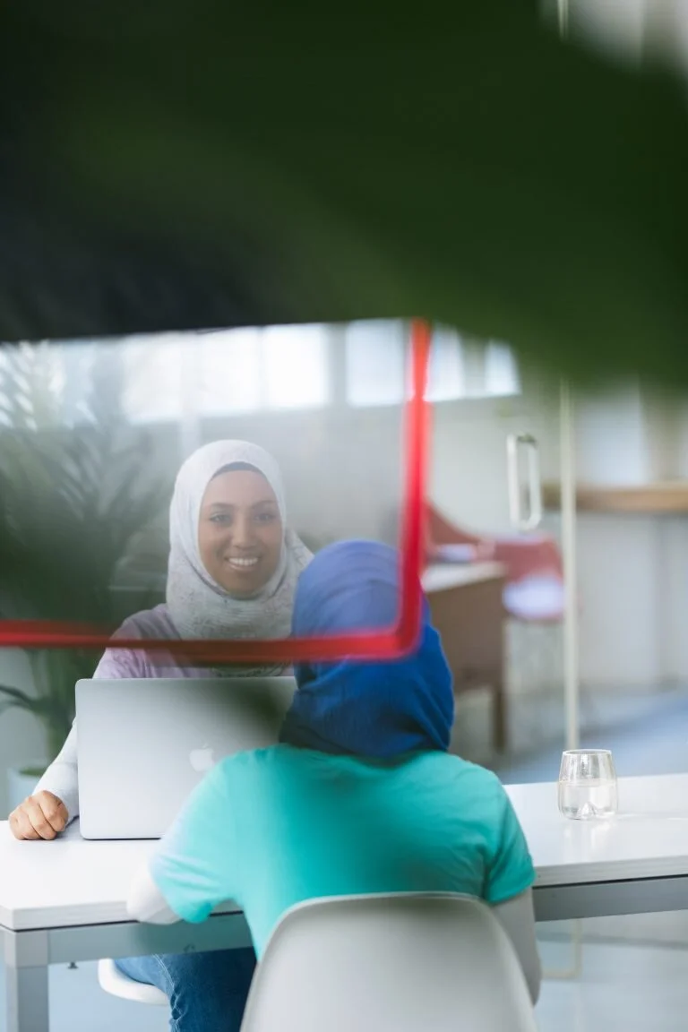 A woman wearing a hijab and a purple shirt sits at a desk with a laptop, smiling and looking at a person in a blue hoodie who is sitting across from her. The image is partially obscured by a large green leaf in the foreground.