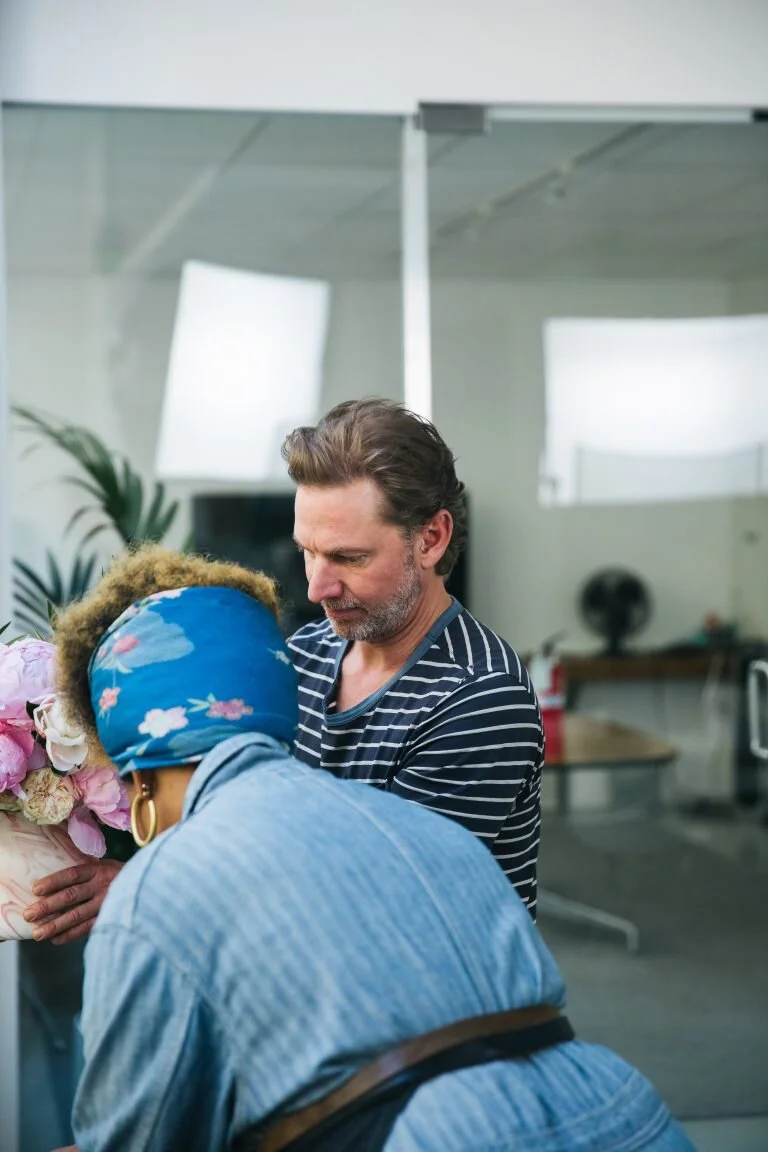 A man and a woman in an indoor setting, with the woman bowing her head towards a bouquet of pink and white flowers, and the man looking at the flowers, in a likely heartfelt moment.