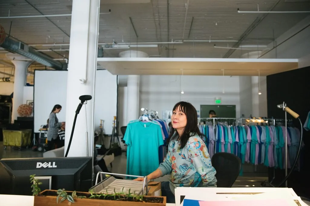 A woman sitting at a desk in an office or retail space with racks of colorful clothes in the background.
