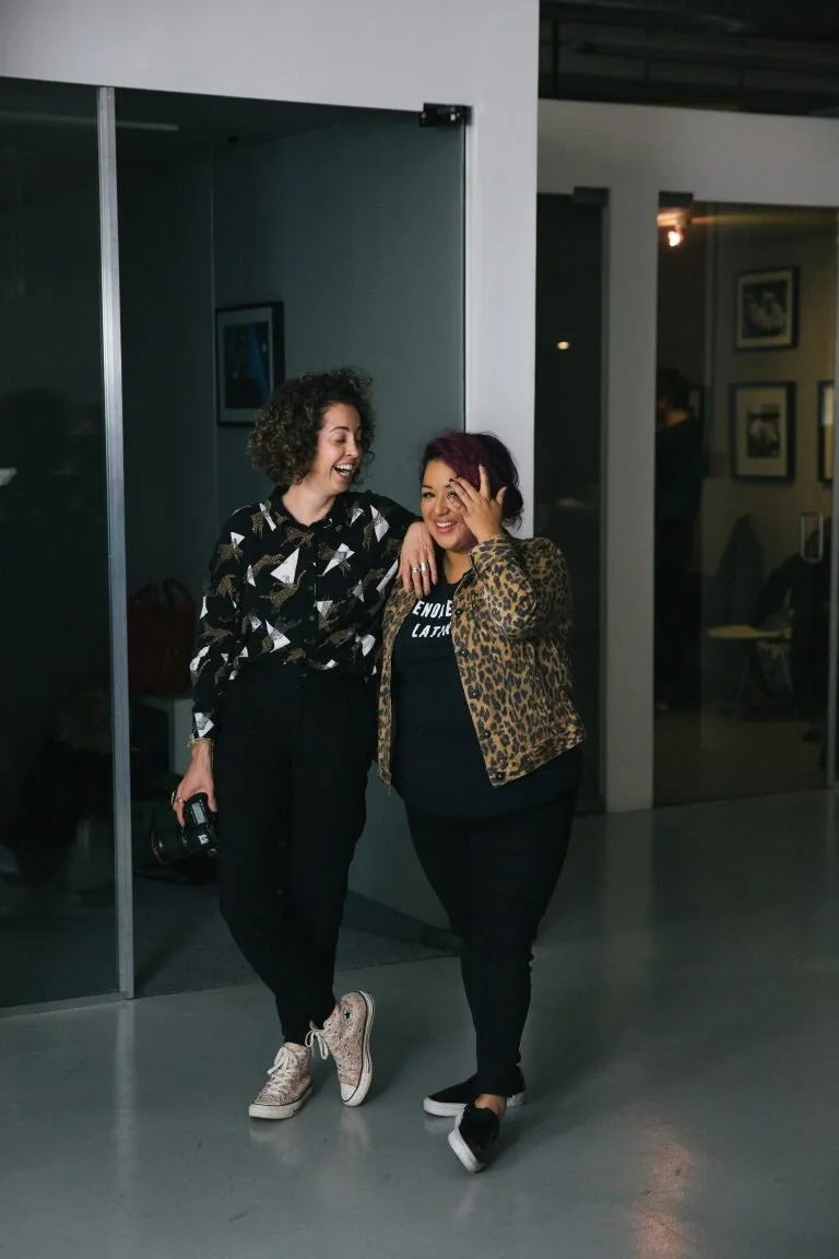 Two women laughing and enjoying each other's company indoors, one wearing a black patterned shirt and sneakers, the other wearing a leopard print jacket and black sneakers.