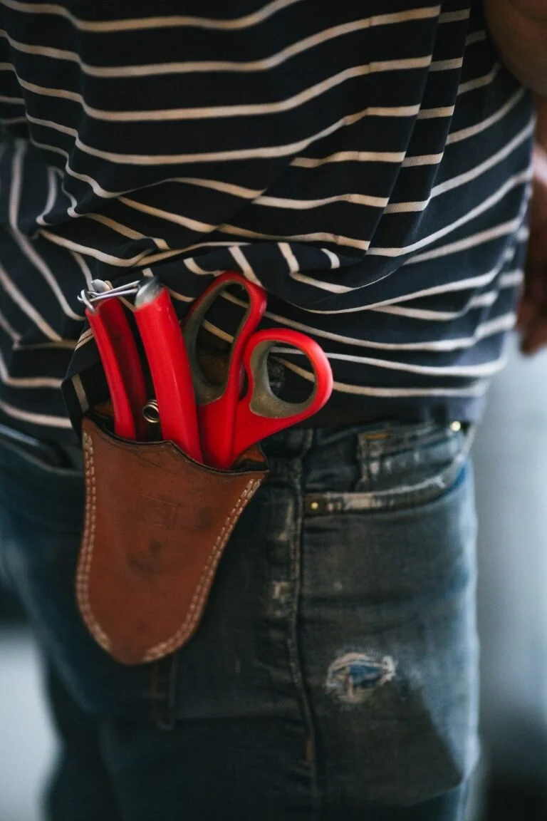 A person wearing a striped shirt and jeans with a brown holster holding red scissors and a red utility knife in the belt.
