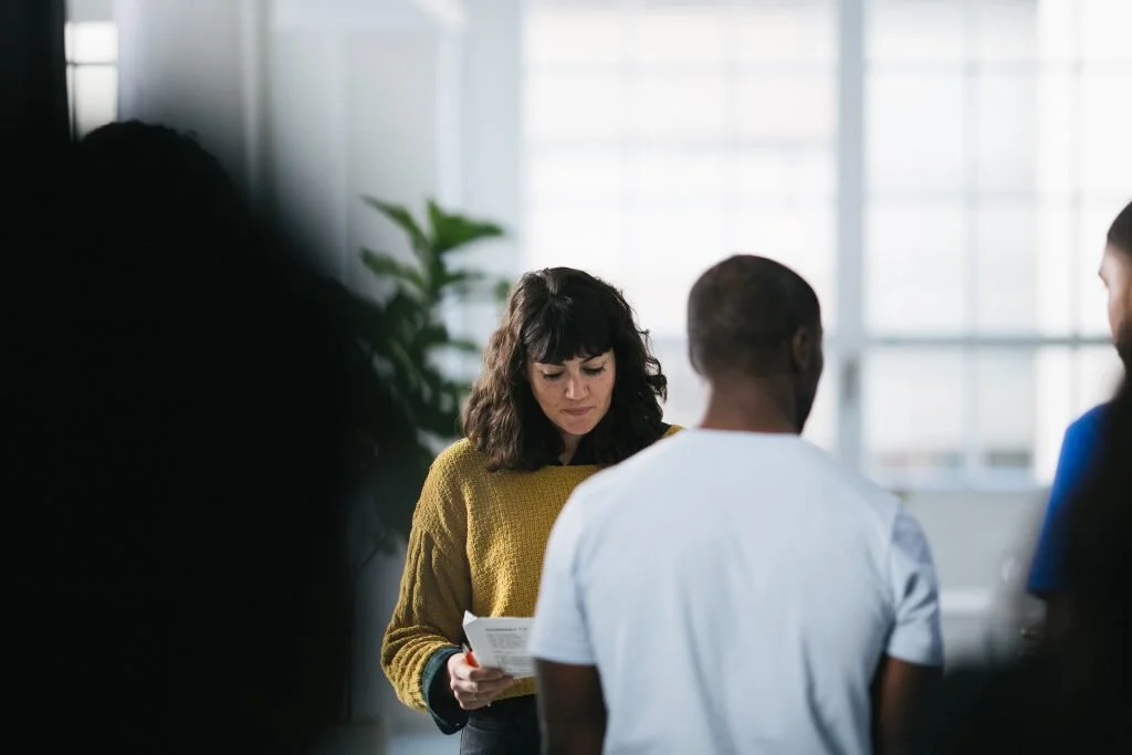 Group of people in a meeting or discussion in a well-lit room with large windows and a potted plant, woman in yellow sweater looking down at papers.