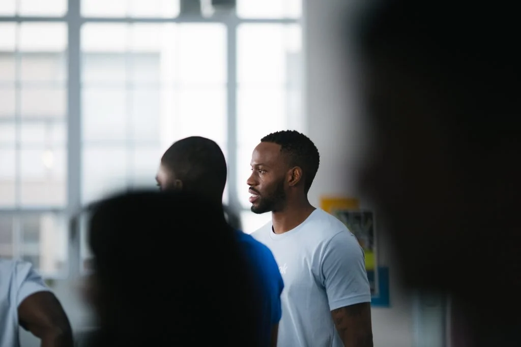 A man with a beard and short hair wearing a light blue T-shirt, standing indoors near large windows, with blurred figures around him.