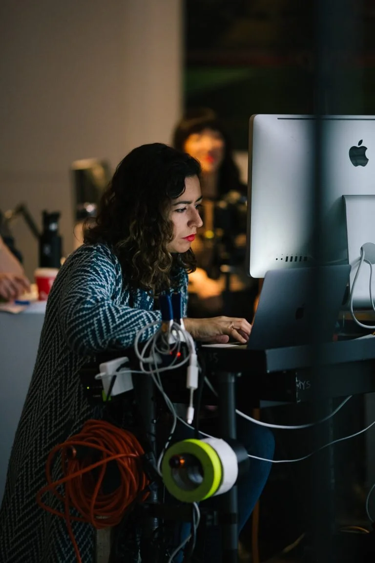 Woman with curly hair working on a desktop computer surrounded by electronic equipment in an office or studio setting.