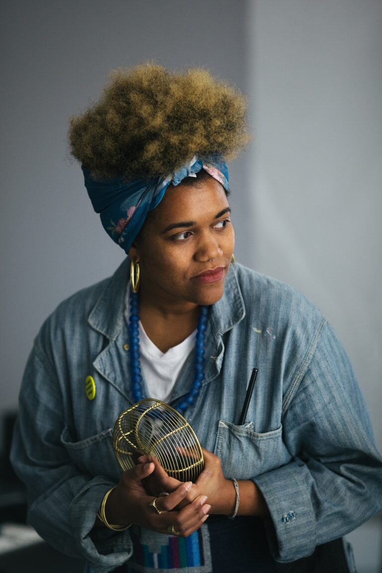A woman with curly blonde hair tied with a colorful headscarf, wearing gold hoop earrings, a denim jacket with a yellow badge, and a blue beaded necklace. She is holding a small golden wire cage and appears to be looking to her right.
