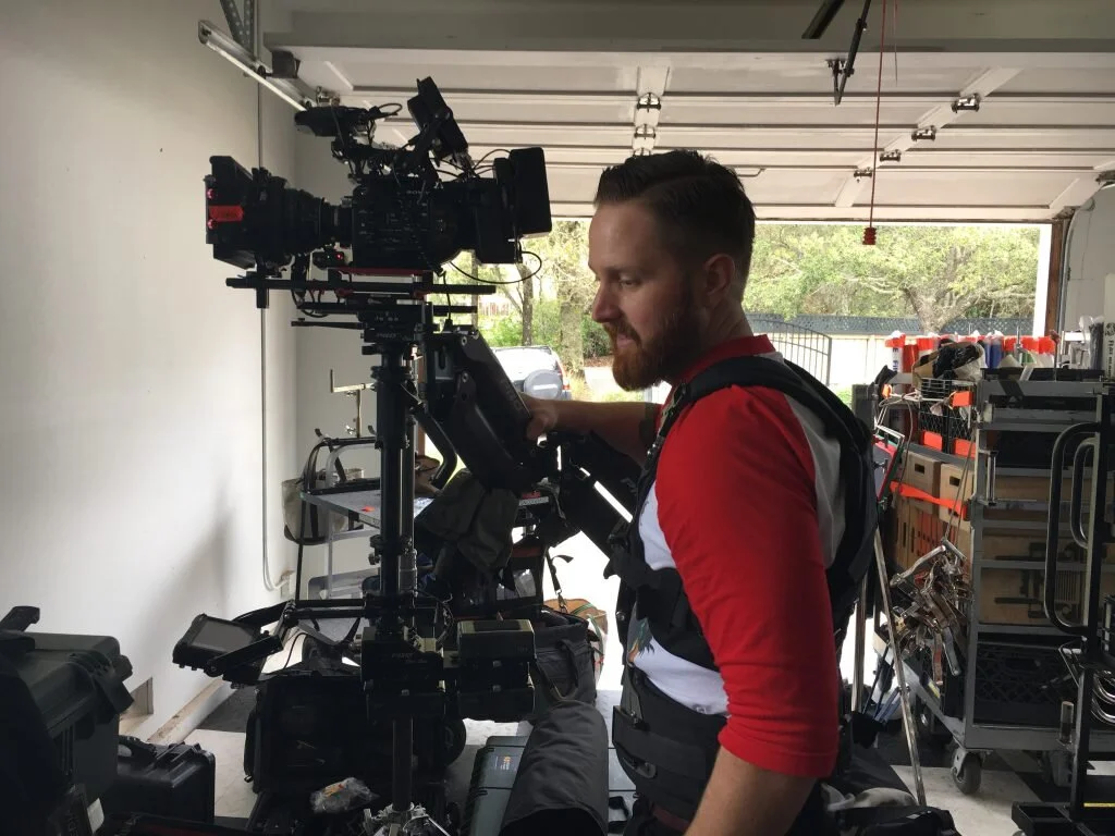 Man in a red, gray, and white shirt working with a camera rig in a garage or workshop setting.