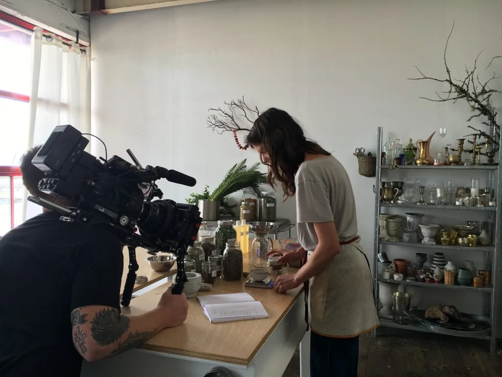 Woman in beige apron preparing ingredients in a kitchen with various jars, plants, and kitchenware on the counter and shelves. Man filming her with a camera.