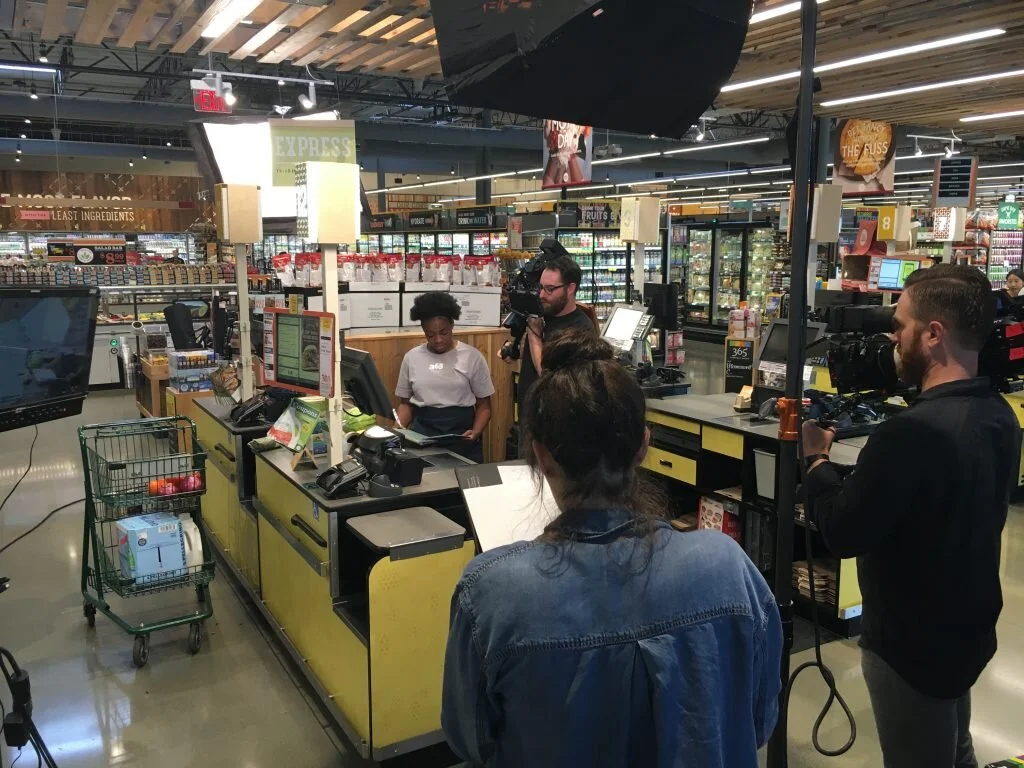 A filming crew in a grocery store checkout area, with a person recording at the checkout counter, and others operating cameras and equipment.