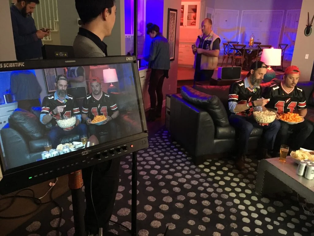 People watching a television screen that shows two men in football jerseys sitting on a couch, eating snacks. Some individuals are seated on the couch and others are standing around in a living room with dim, colorful lighting, and decorated walls.