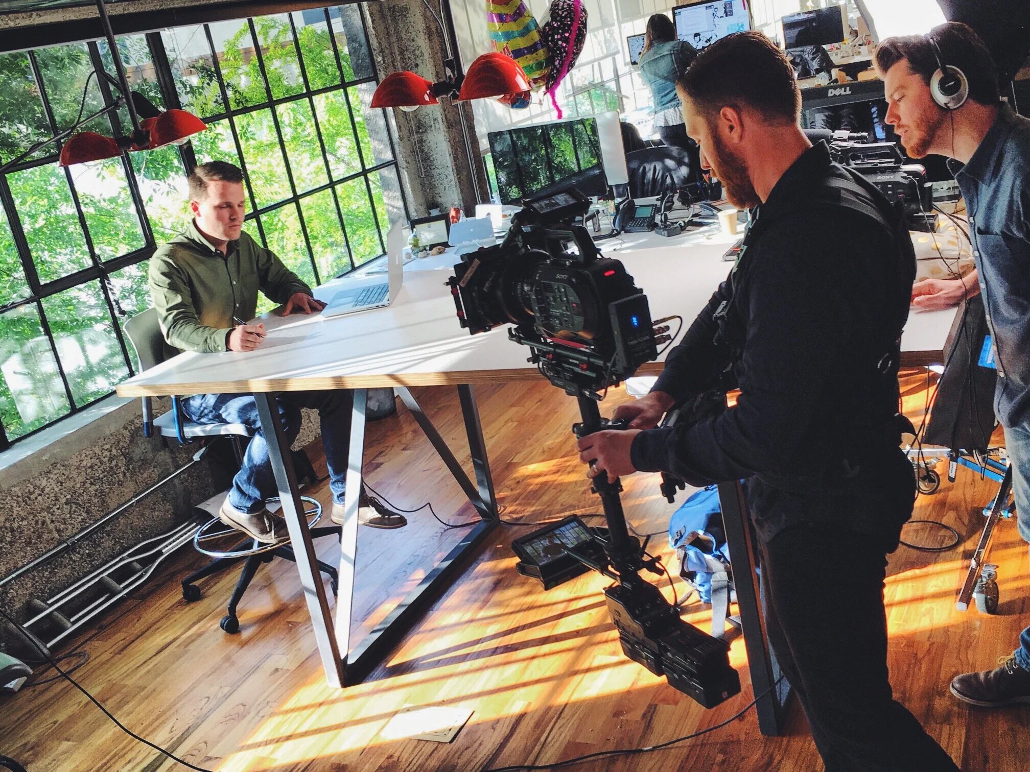 Three men working on a video shoot in a modern office space with large windows, greenery outside, and multiple computer monitors. One man is holding a professional camera, and another is wearing a headset.