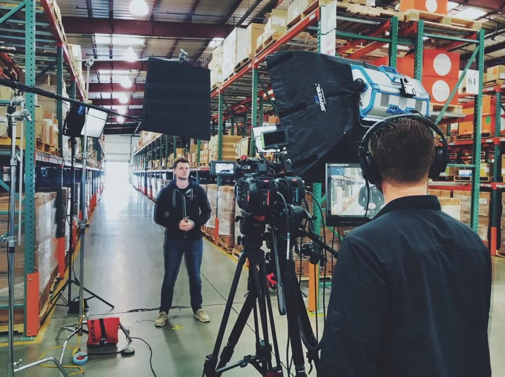 A man being filmed in a warehouse aisle with shelves stocked with boxes, using video equipment including a camera, monitor, lights, and a crew member wearing headphones.