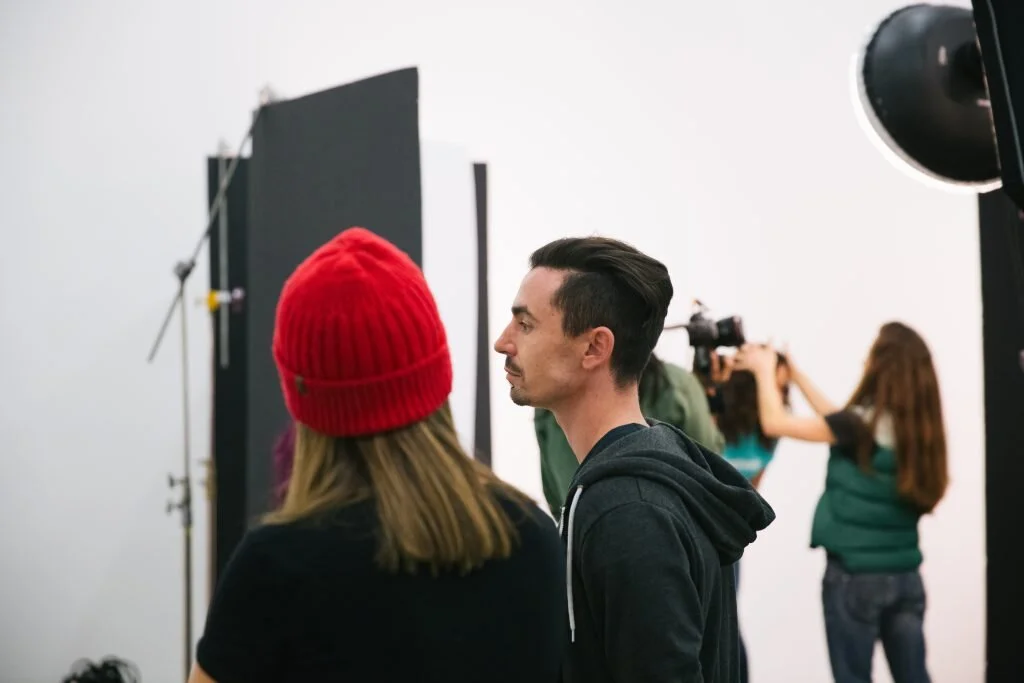 People on a film set with a backdrop, camera, and lighting equipment. One person is wearing a red beanie, and another has dark, slicked-back hair and a black hoodie.