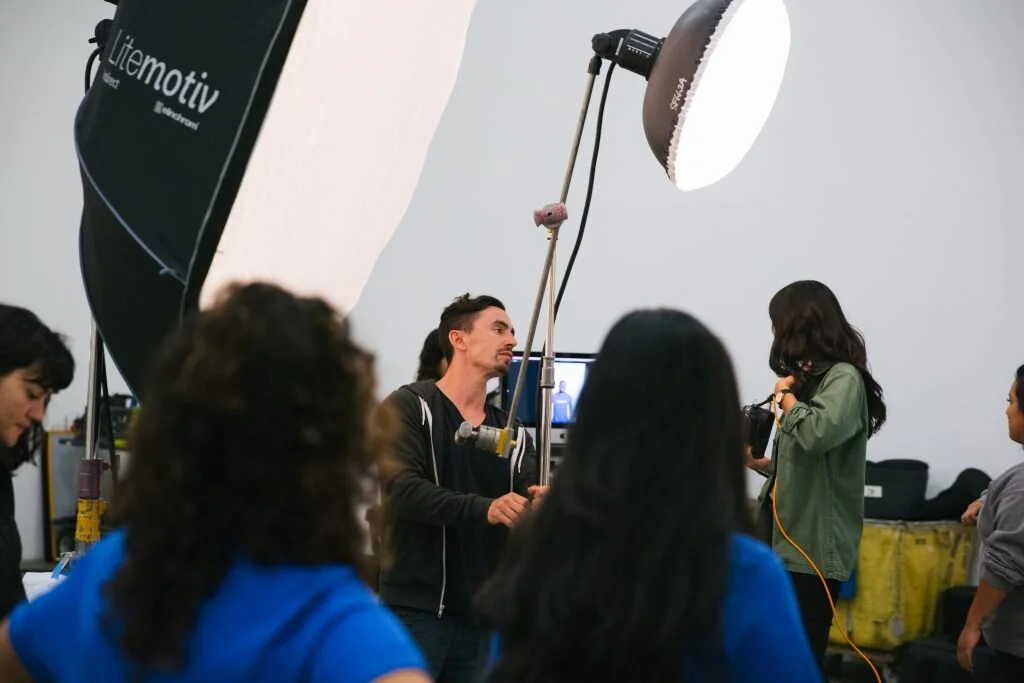 A group of people behind the scenes on a film or photography set, with a large lighting softbox overhead and a person holding a boom microphone.