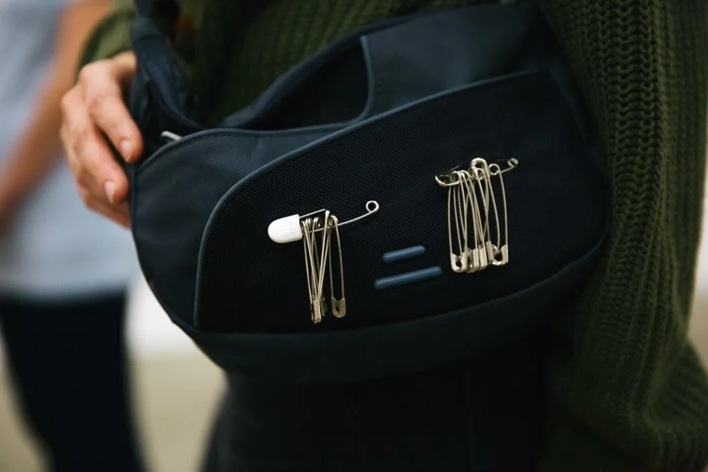 A person with a green sweater holding a black bag with safety pins attached to the front pocket.