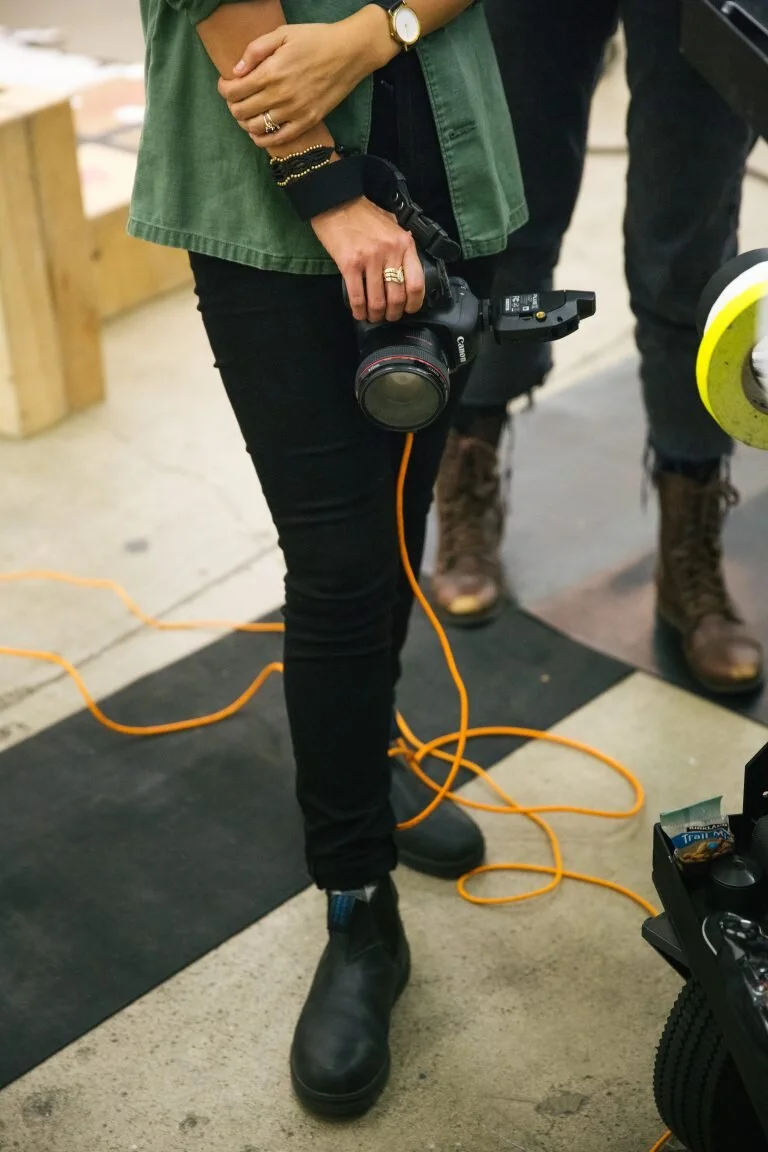 Person holding a Canon camera with a strap, wearing black pants, black boots, and a green jacket, standing on a black mat with orange extension cords on the floor in an indoor setting.