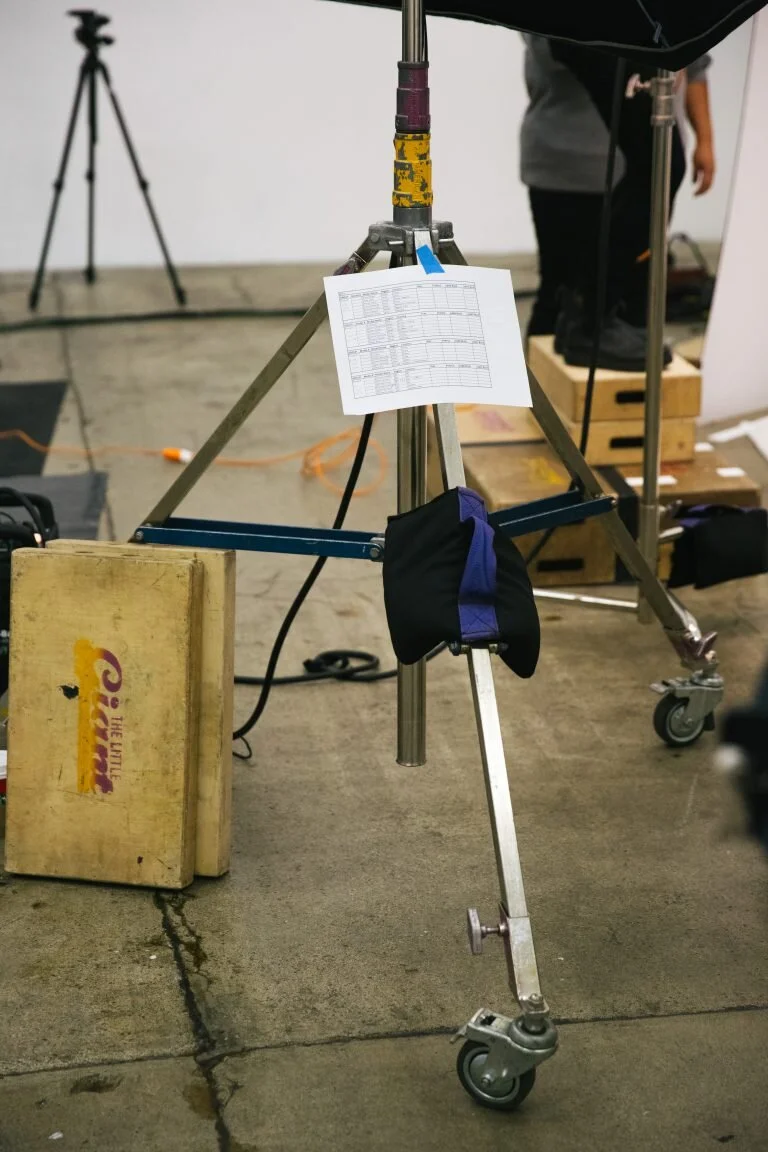 A film or photography set with a tripod holding a white sheet of paper, a wooden box, and various equipment, with a person in the background.