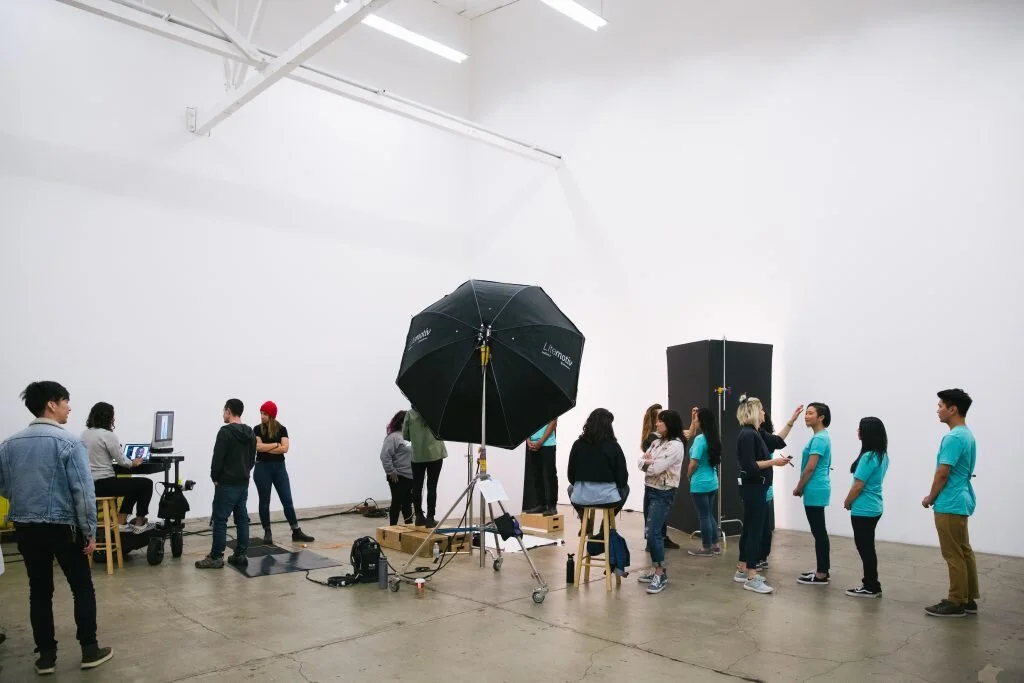 People in a photography or filming studio with lighting equipment, a large umbrella reflector, and a computer workstation. Some individuals are working with the camera, while others are waiting or talking.