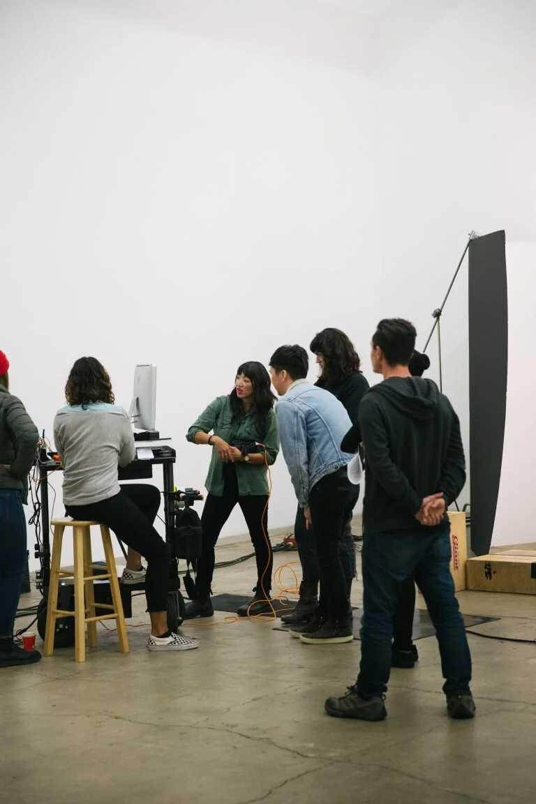 A group of people in a studio, with one woman standing and talking to others, surrounded by photography equipment and a white backdrop.
